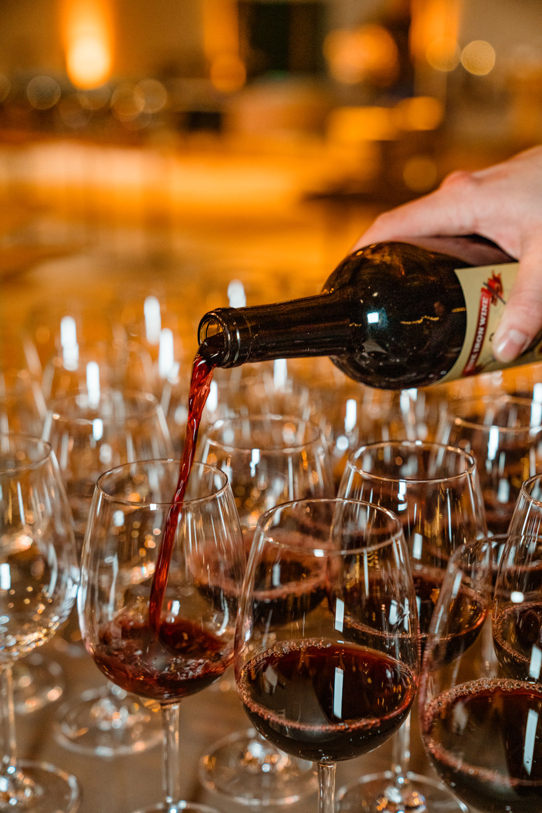 Red wine poured from a bottle into rows of stemmed glasses at the hangar bar