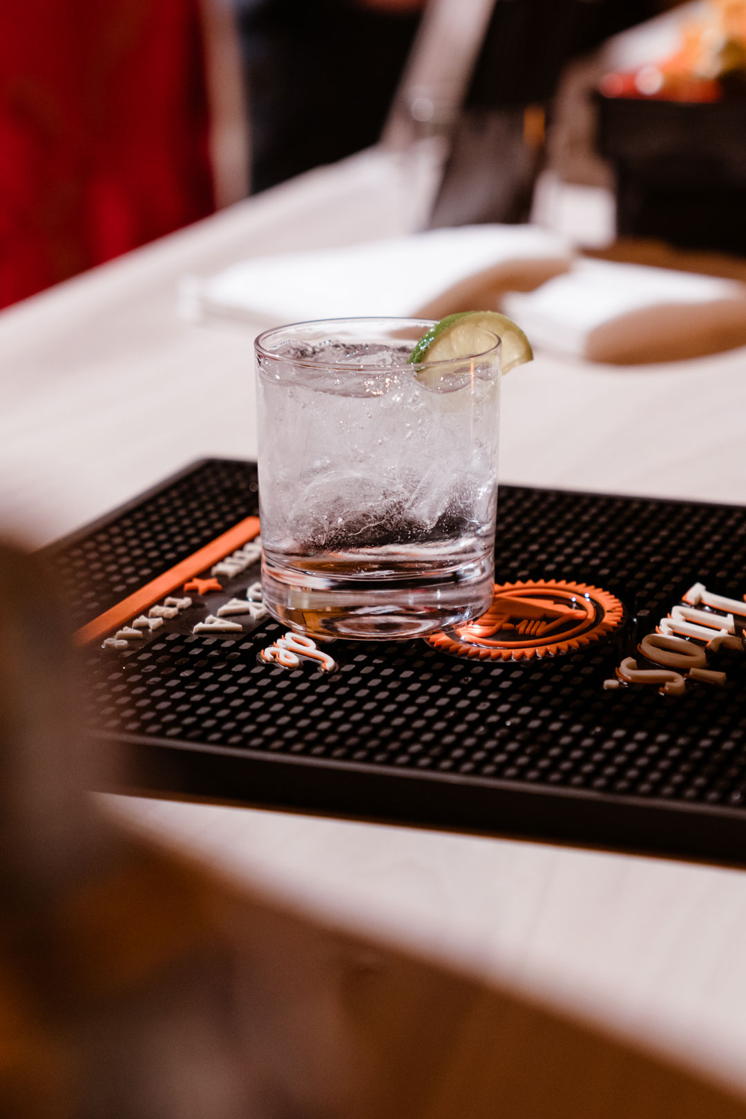 Clear cocktail over ice with a lime wedge in a rocks glass on a black rubber bar mat
