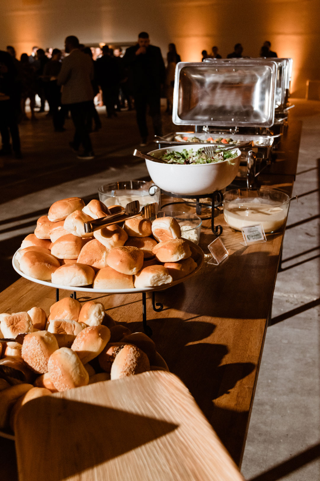 Dinner buffet with platters of fresh baked rolls, a salad bowl and chafing dishes, guests mingling behind