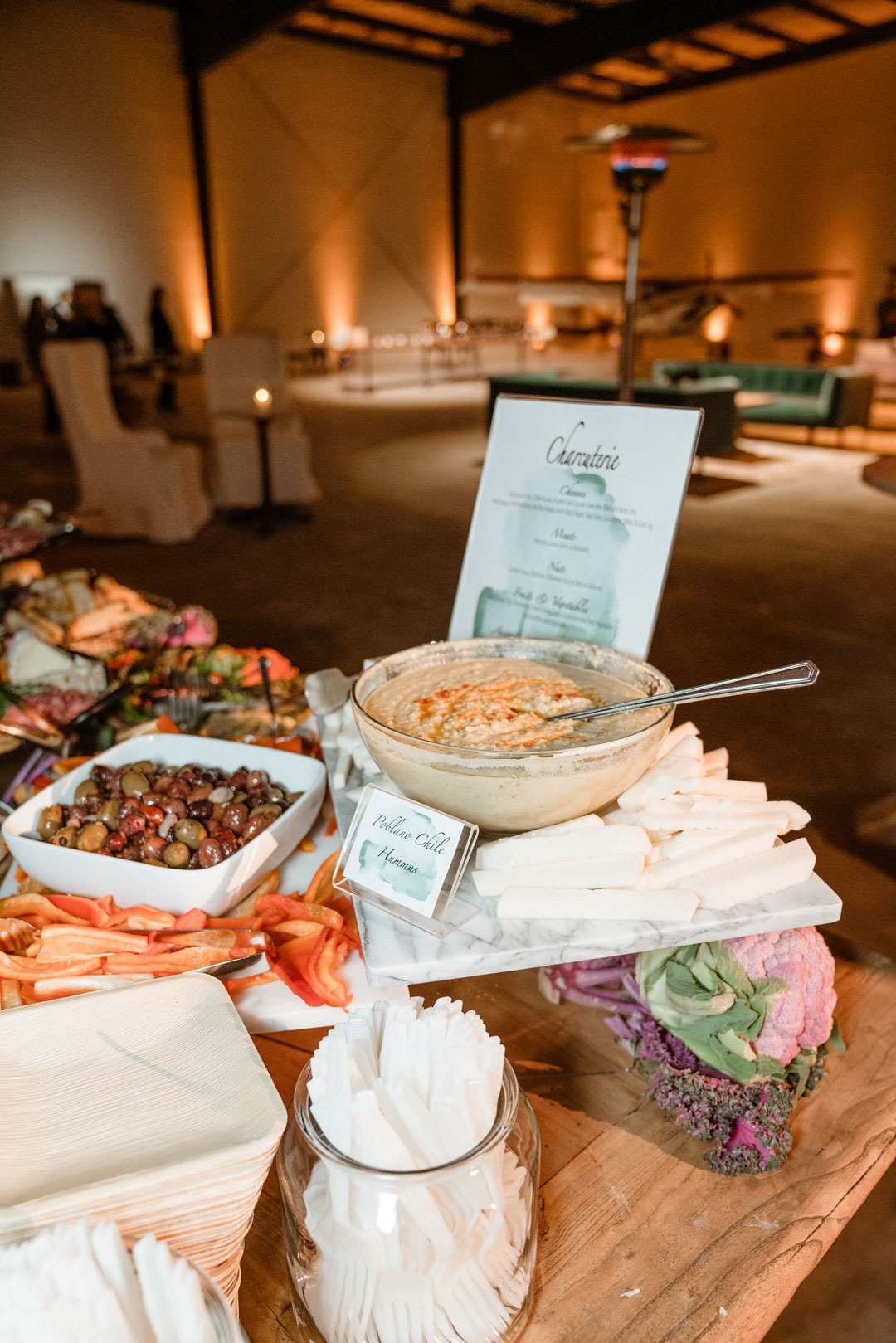 Hummus bowl with vegetable sticks and olives on a marble slab, charcuterie menu card and hangar lounge behind