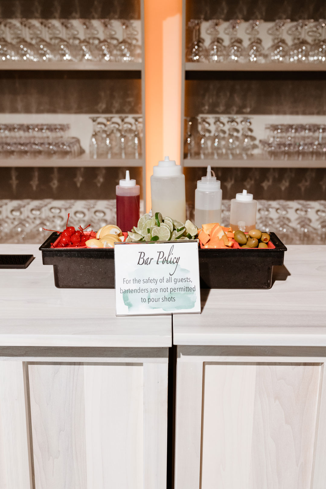 Bar garnish tray of cherries, limes, oranges and olives beside a Bar Policy sign and glassware shelves