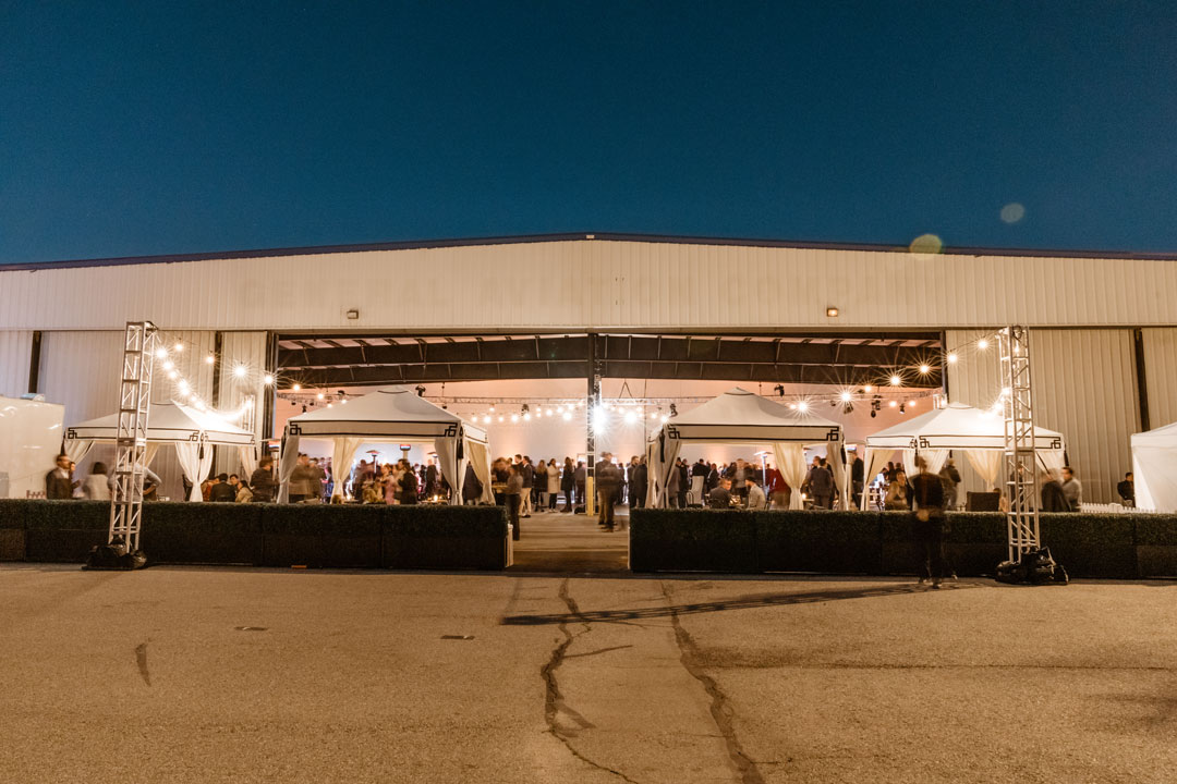 Exterior of The Executive Hangar at dusk with open door, white cabanas, string lights and guests on the tarmac