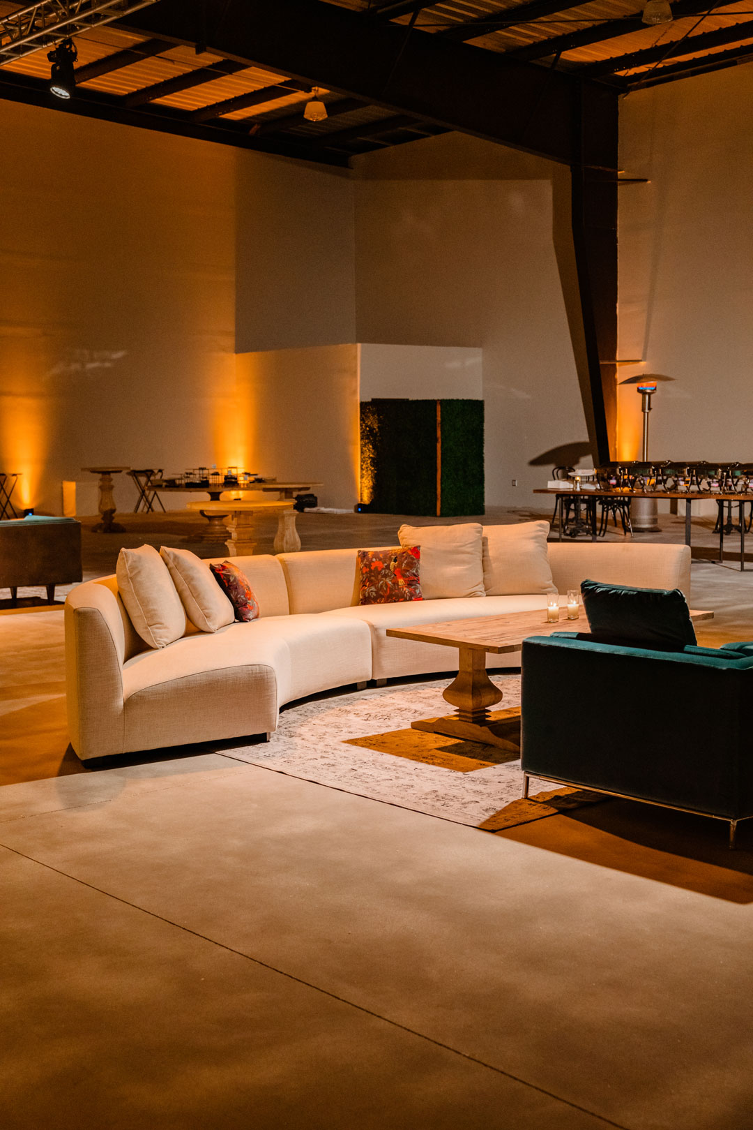Curved cream sectional sofa with patterned pillows and a teal armchair on a rug inside the amber-lit hangar