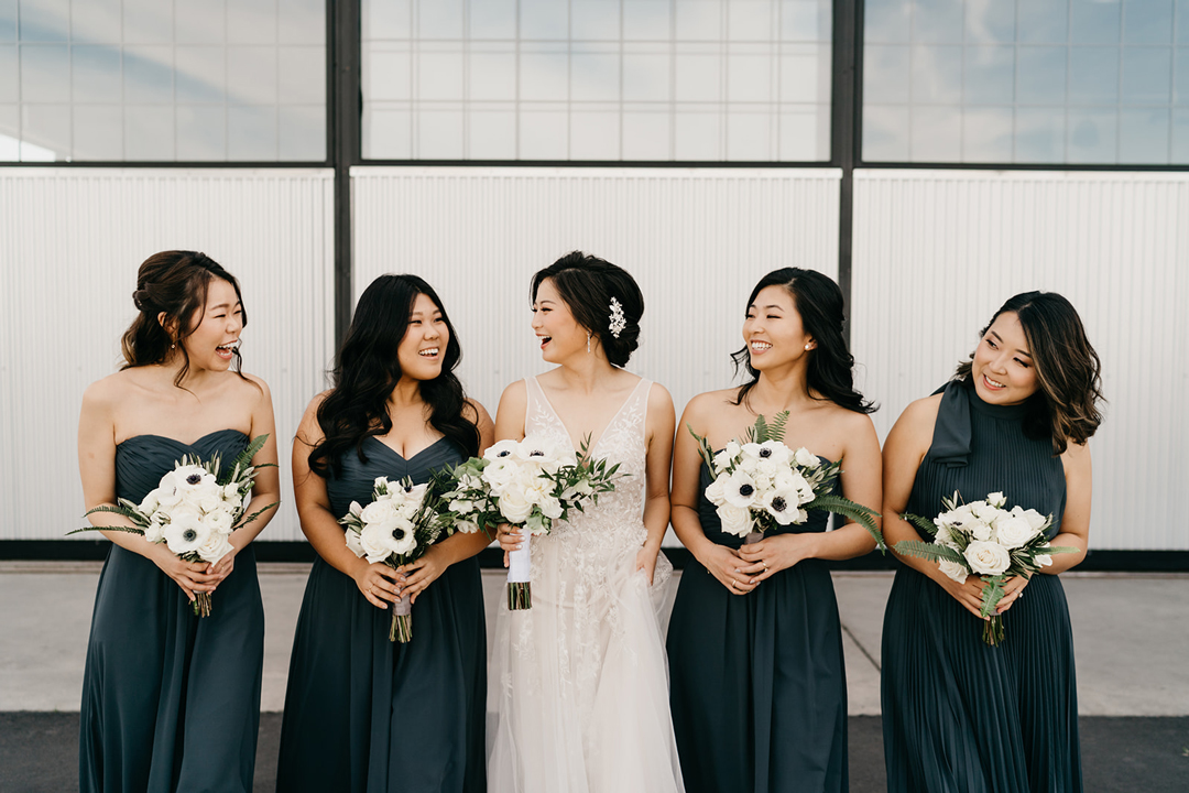 Kayla with her four bridesmaids in dark green gowns holding white bouquets outside Hangar 21 South