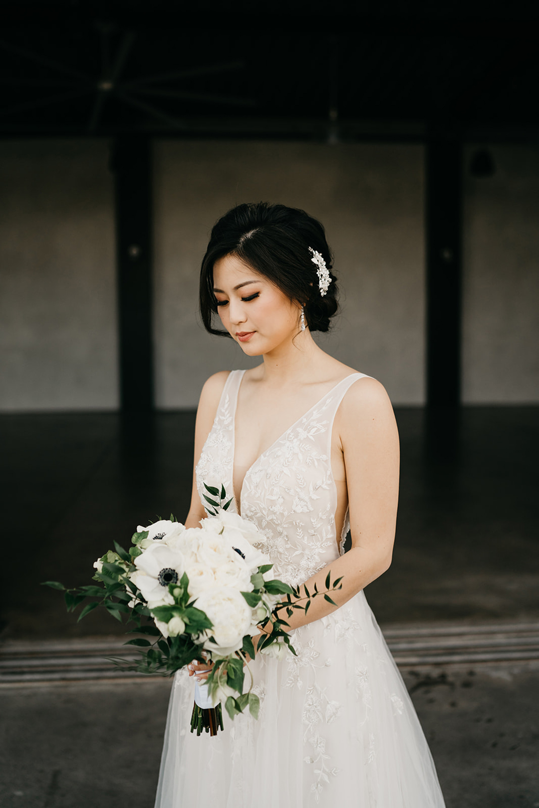 Kayla in a lace V-neck gown looking down at her white anemone bouquet inside the hangar before the first look