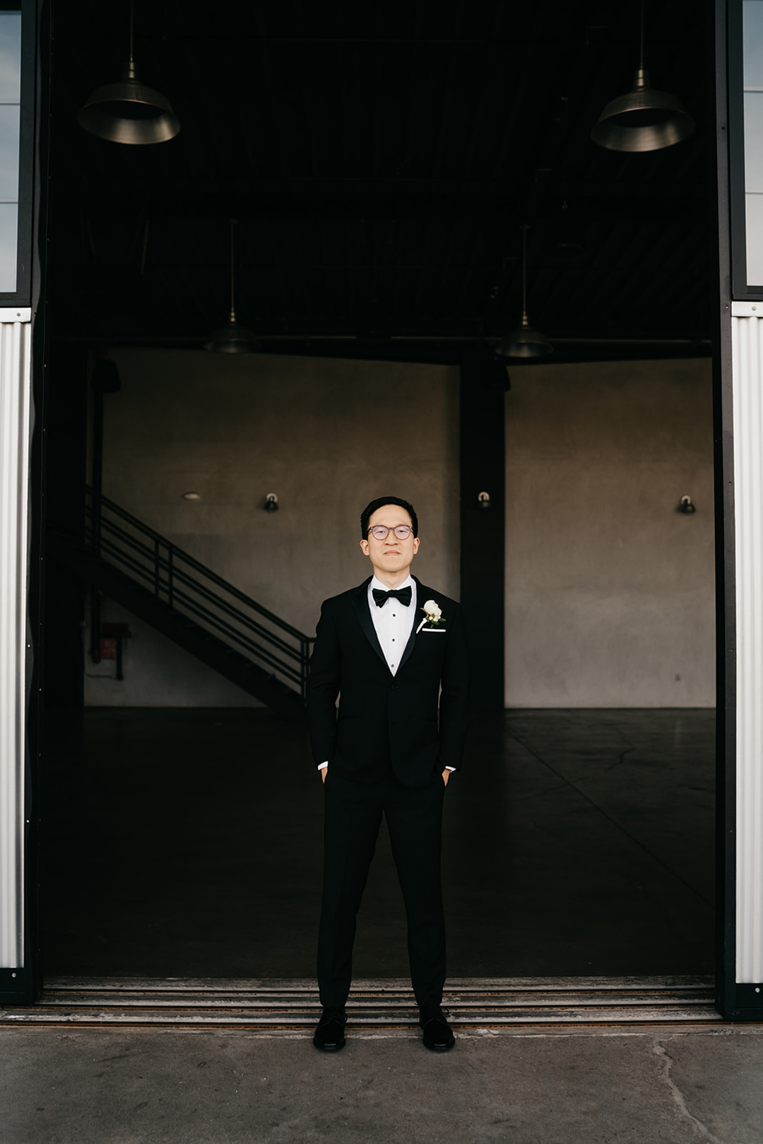 Justin in a black tuxedo and bow tie standing in the open hangar doorway with hands in his pockets before the wedding