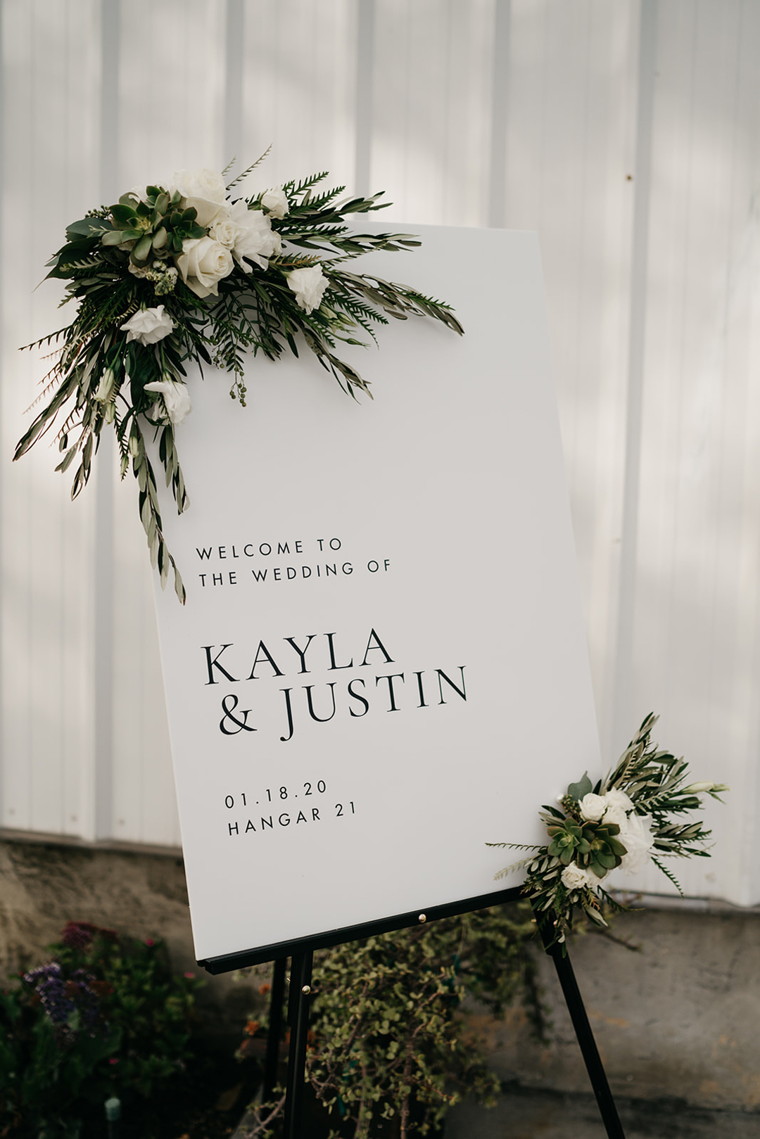 White acrylic welcome sign for Kayla and Justin decorated with white roses, succulents and greenery on an easel