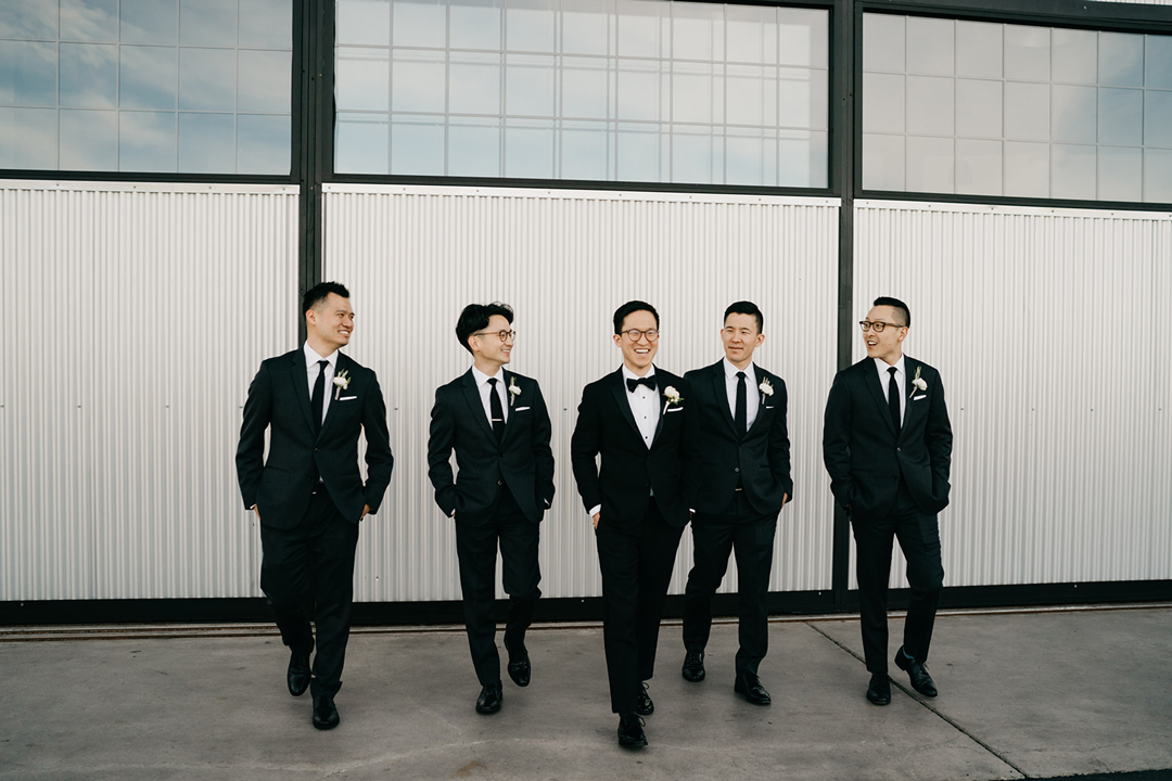 Justin and his groomsmen in black suits walking and laughing along the corrugated hangar wall at Hangar 21