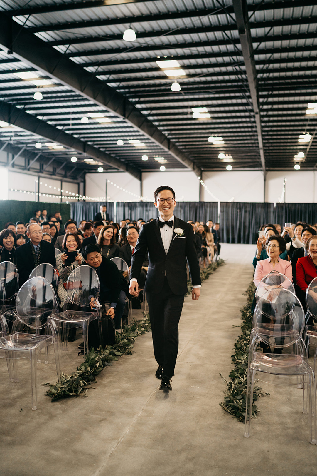 Justin walking down the greenery-lined ceremony aisle under the steel hangar ceiling as guests smile