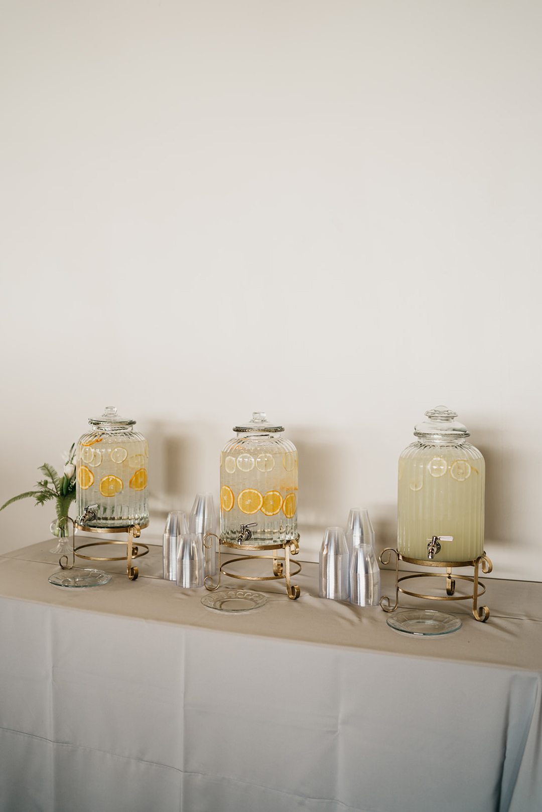 Three glass beverage dispensers with citrus-infused water and lemonade on a grey linen table at cocktail hour