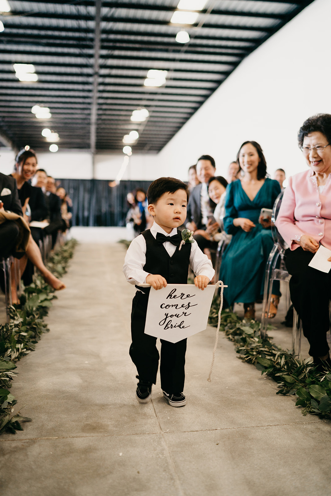 Young ring bearer in a vest and bow tie carrying a here comes your bride banner down the hangar aisle