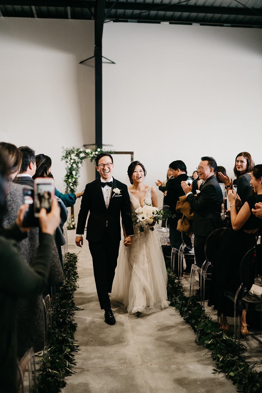 Kayla and Justin laughing as they recess down the aisle past clapping guests seated in clear ghost chairs