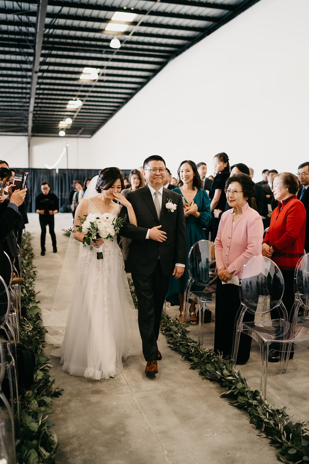 Kayla arm in arm with her father walking down the garland-lined aisle at The Executive Hangar as guests look on