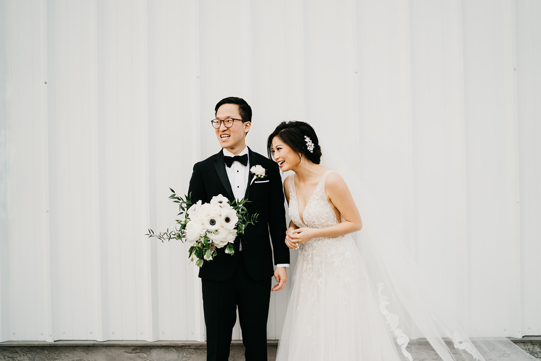 Newlyweds Kayla and Justin laughing together with her white bouquet against the hangar's white metal wall
