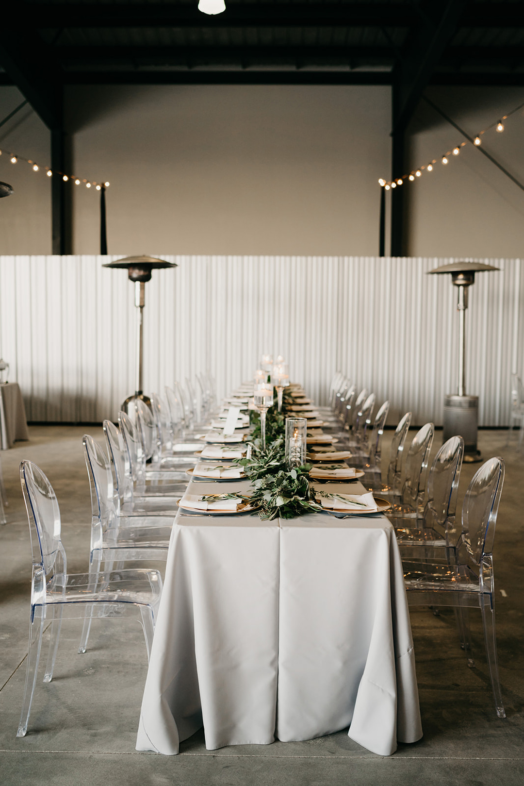Long reception table with grey linen, gold chargers, greenery garland and clear ghost chairs inside the hangar