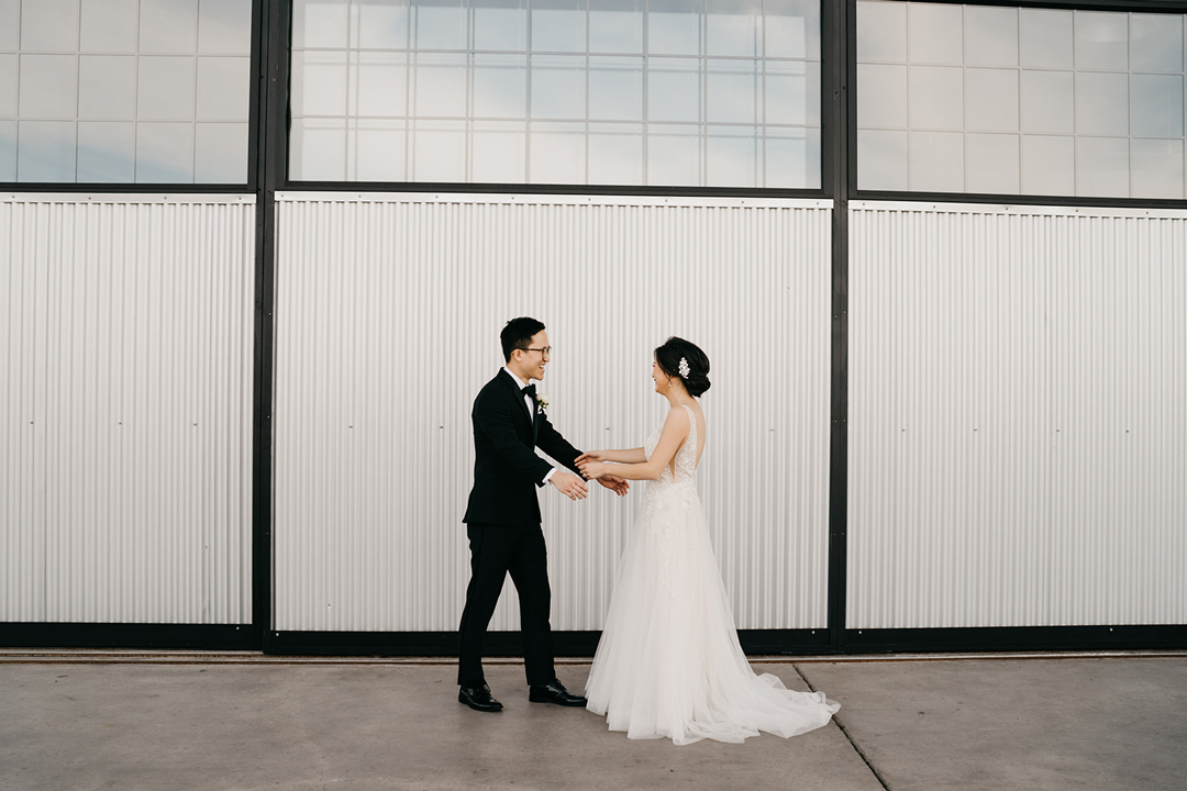 Kayla and Justin holding hands and laughing after their first look against the white paneled hangar exterior