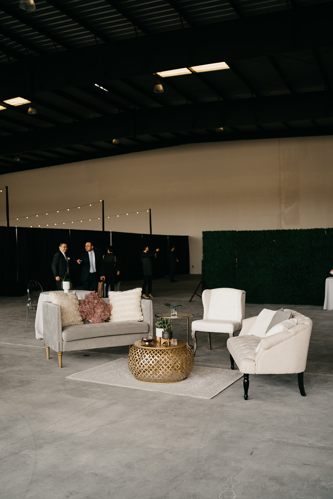 Cocktail lounge seating with plush sofas, a gold drum table and boxwood backdrop in the open hangar