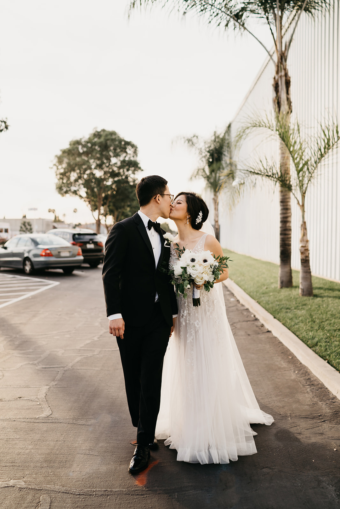 Kayla and Justin sharing a kiss while walking beside palm trees outside the hangar at golden hour