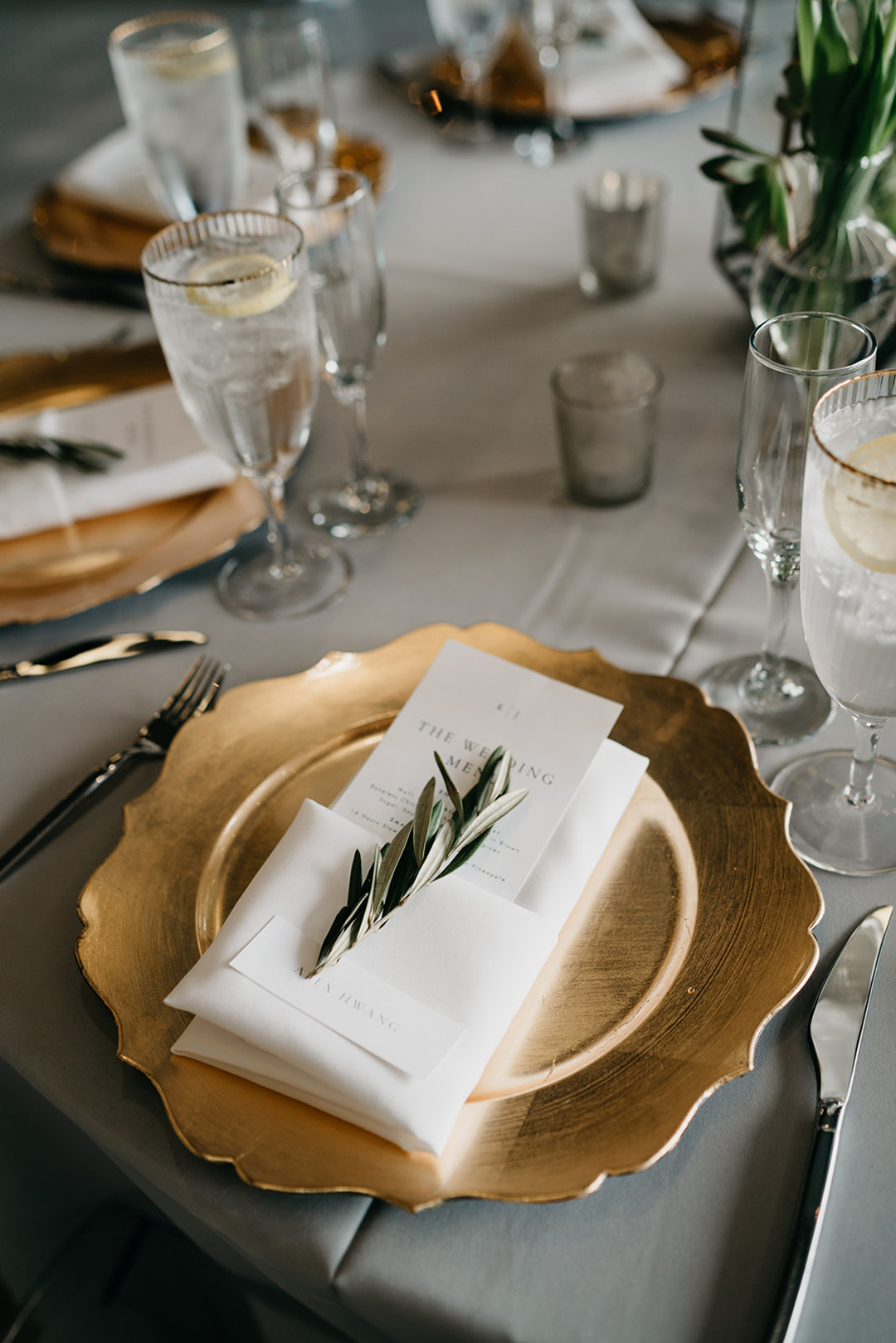 Gold scalloped charger plate with a menu card, folded napkin and olive sprig on a grey linen reception table