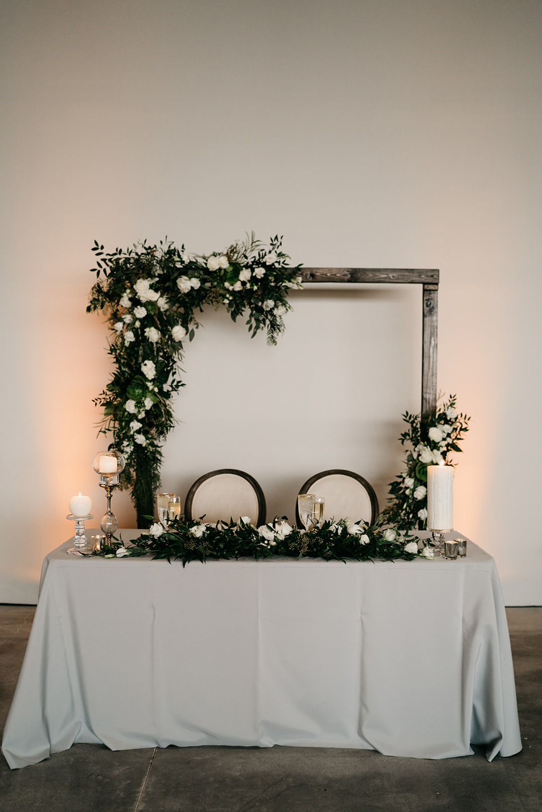 Sweetheart table with a wooden floral frame backdrop, greenery garland and candles on grey linen