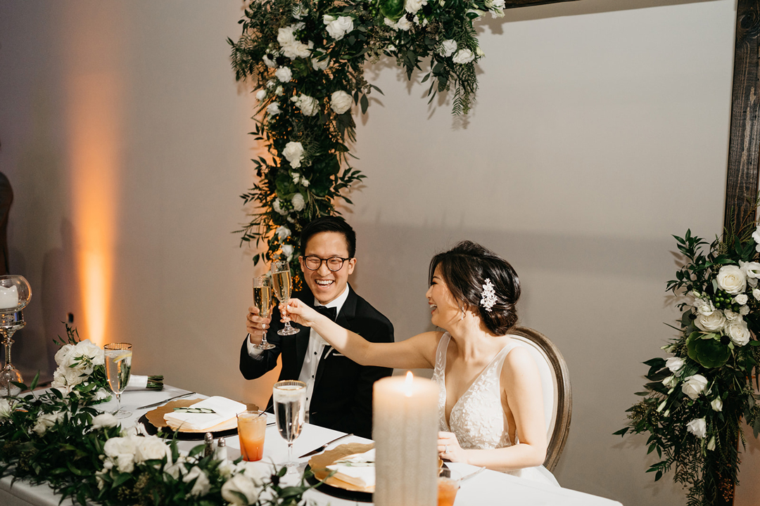 Kayla and Justin toasting with champagne at the flower-framed sweetheart table during reception speeches