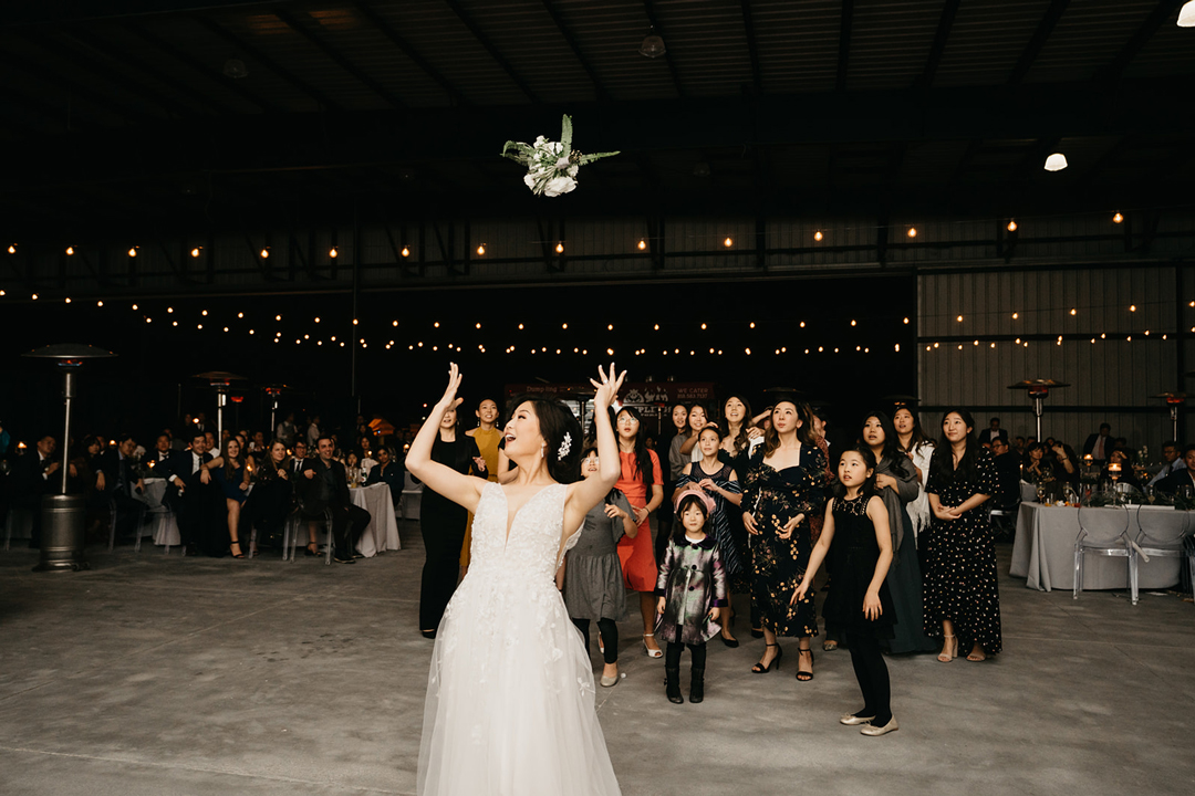 Kayla tossing her bouquet to a crowd of eager guests under string lights inside the hangar