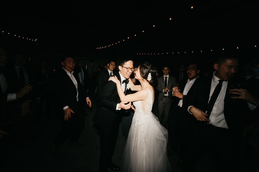 Kayla and Justin dancing surrounded by groomsmen on the dance floor under dim string lights