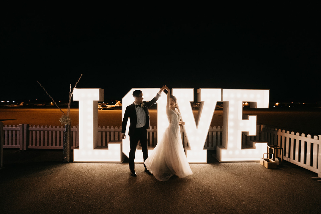 Kayla and Justin dancing in front of giant illuminated LOVE marquee letters on the airport tarmac at night