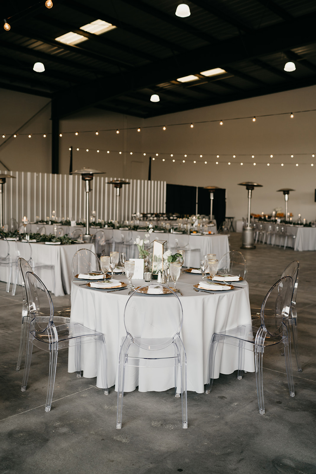 Round reception table with white linen, gold chargers and clear chairs under string lights in the hangar