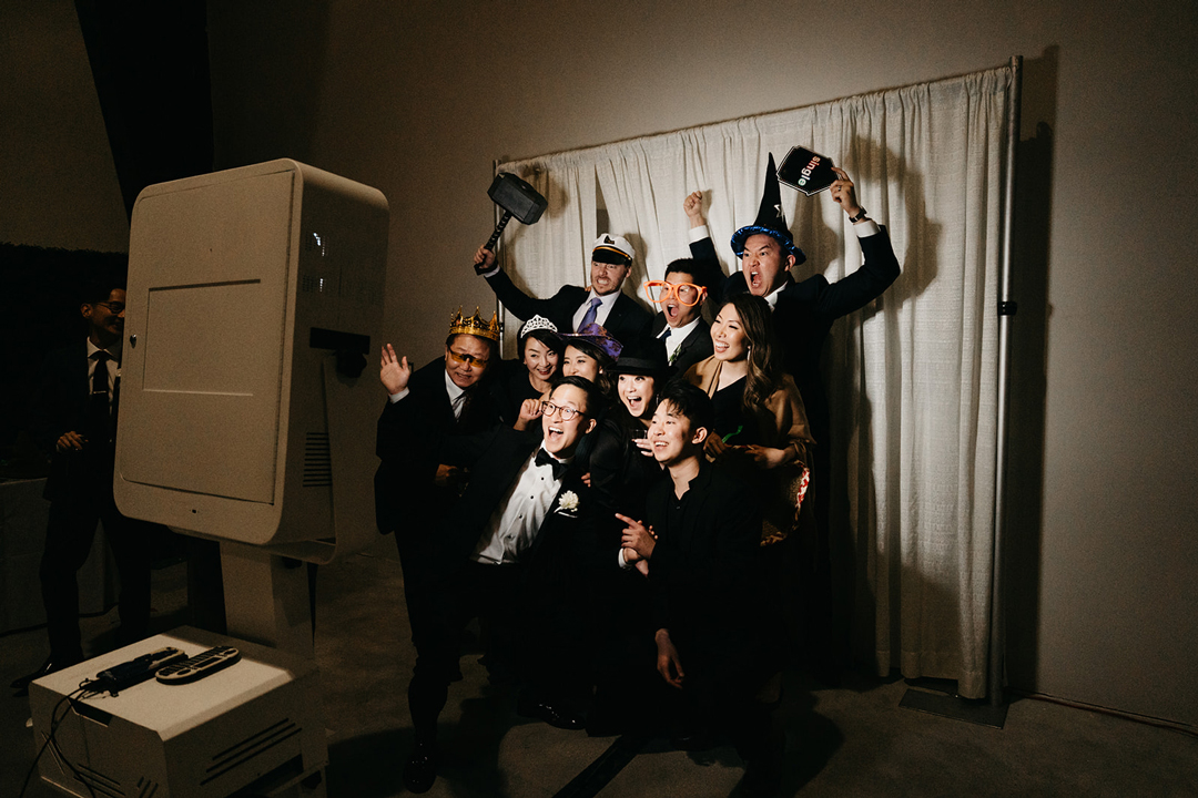 Justin and friends in party hats, crowns and oversized glasses posing in front of the photo booth at the reception