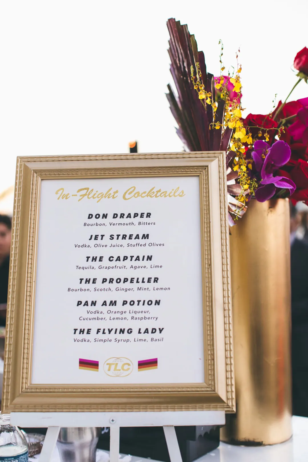 Gold-framed In-Flight Cocktails menu with signature drinks beside a gold vase of orchids and roses at the gala