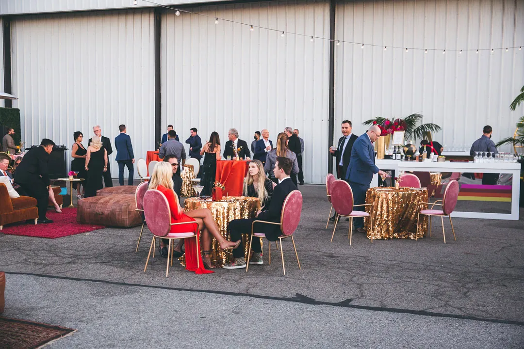 Gala guests seated at gold sequin cocktail tables with pink velvet chairs outside the hangar doors at The Executive Hangar