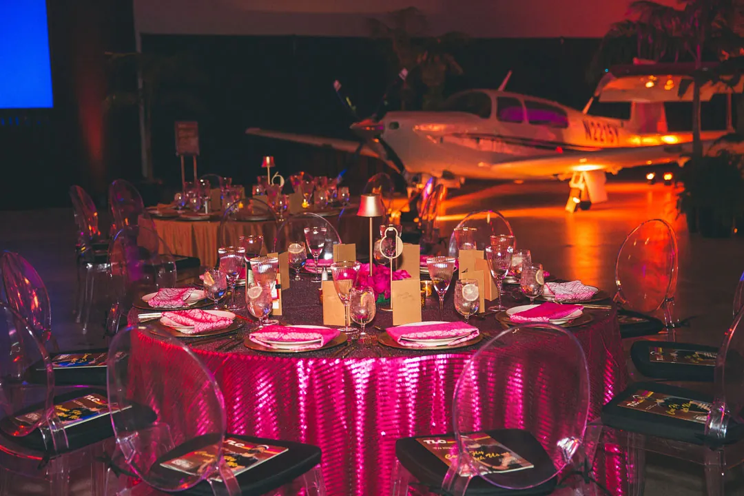 Pink sequin dinner table with ghost chairs set in front of a small plane inside The Executive Hangar