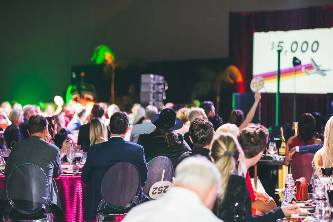 Guests raise a TLC paddle to bid as a $5,000 auction slide shows on screen inside The Executive Hangar