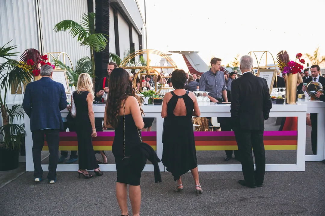 Guests in black attire order drinks at a white bar with Pan Am stripes and gold arches at The Executive Hangar