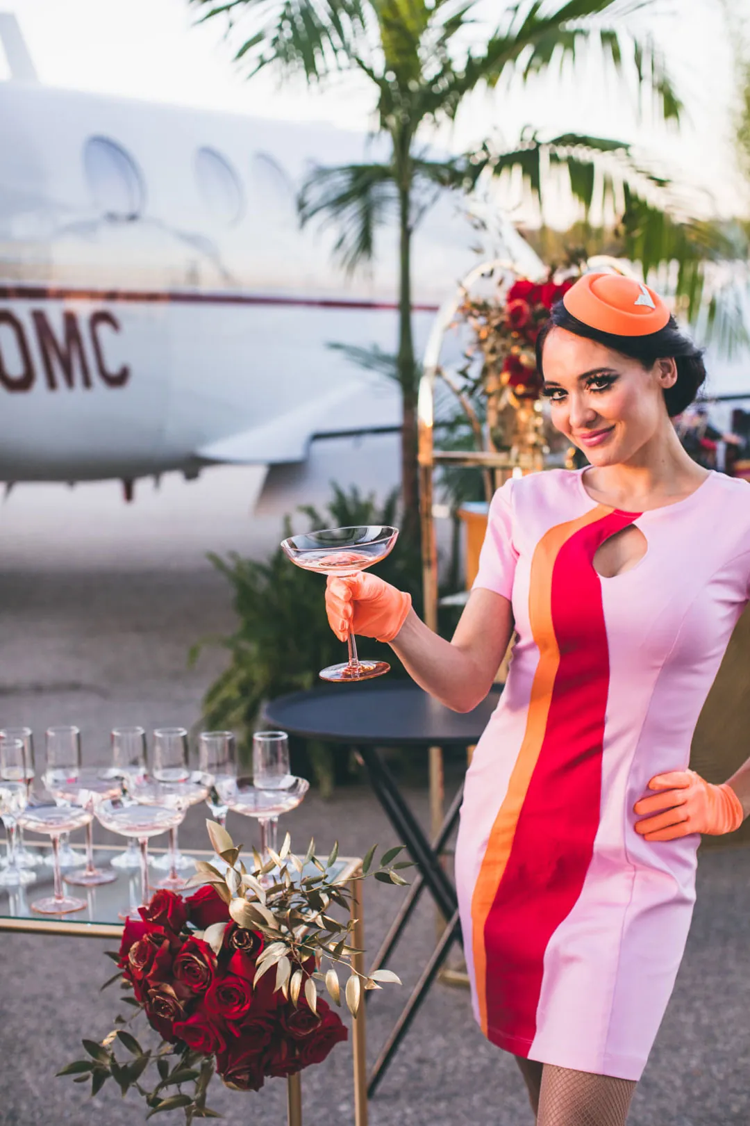 Flight attendant character in a pink and orange retro dress holds a coupe glass beside a jet at The Executive Hangar
