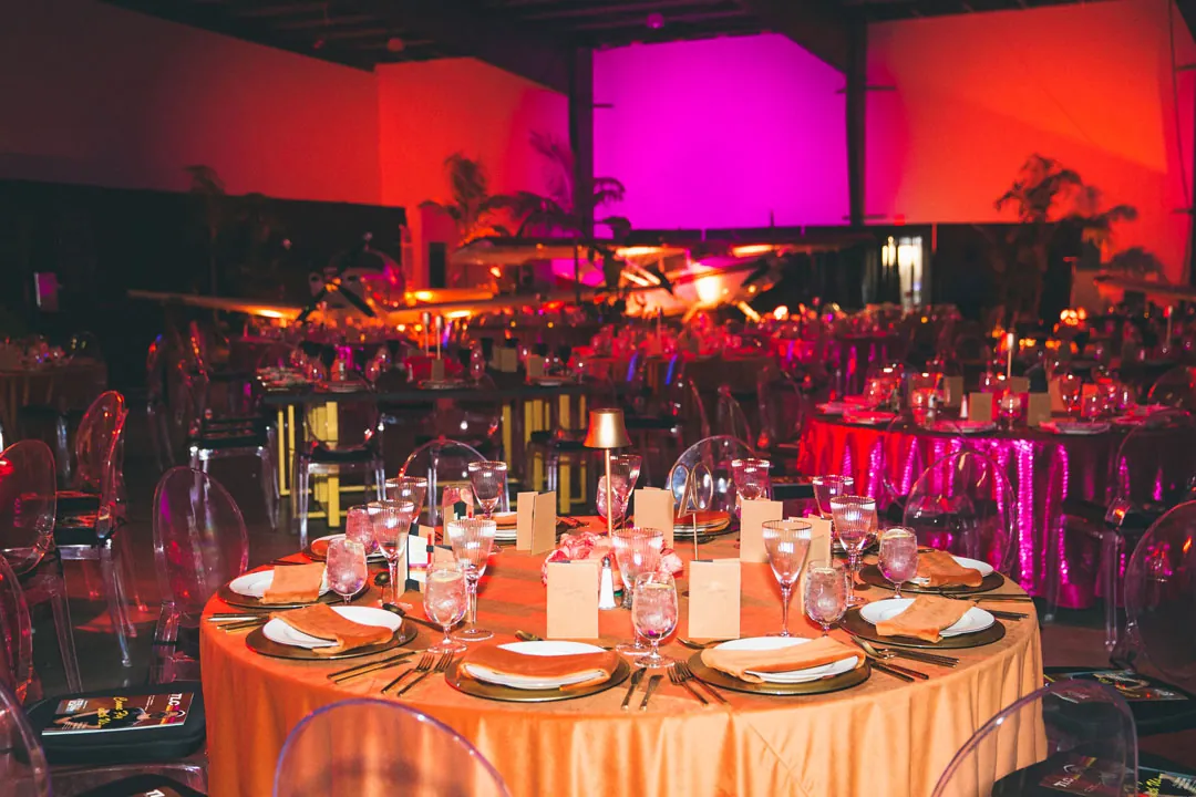 Orange draped dinner table with planes and fuchsia uplighting filling The Executive Hangar behind it