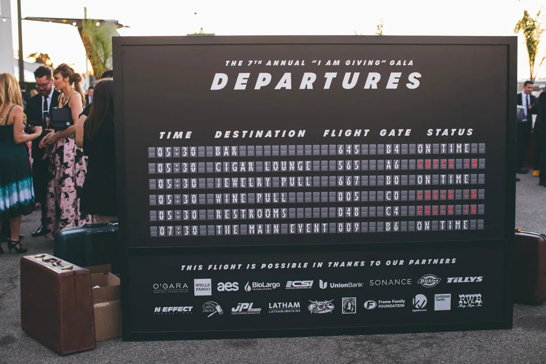 Airport-style Departures board listing bar, cigar lounge and wine pull for the I Am Giving Gala at The Executive Hangar