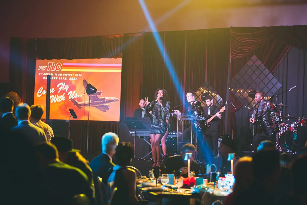 Live band with a singer performs on stage beside a Come Fly With Us screen at the I Am Giving Gala