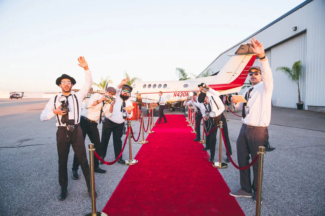 Costumed paparazzi with vintage cameras line a red carpet on the tarmac beside a small plane at The Executive Hangar