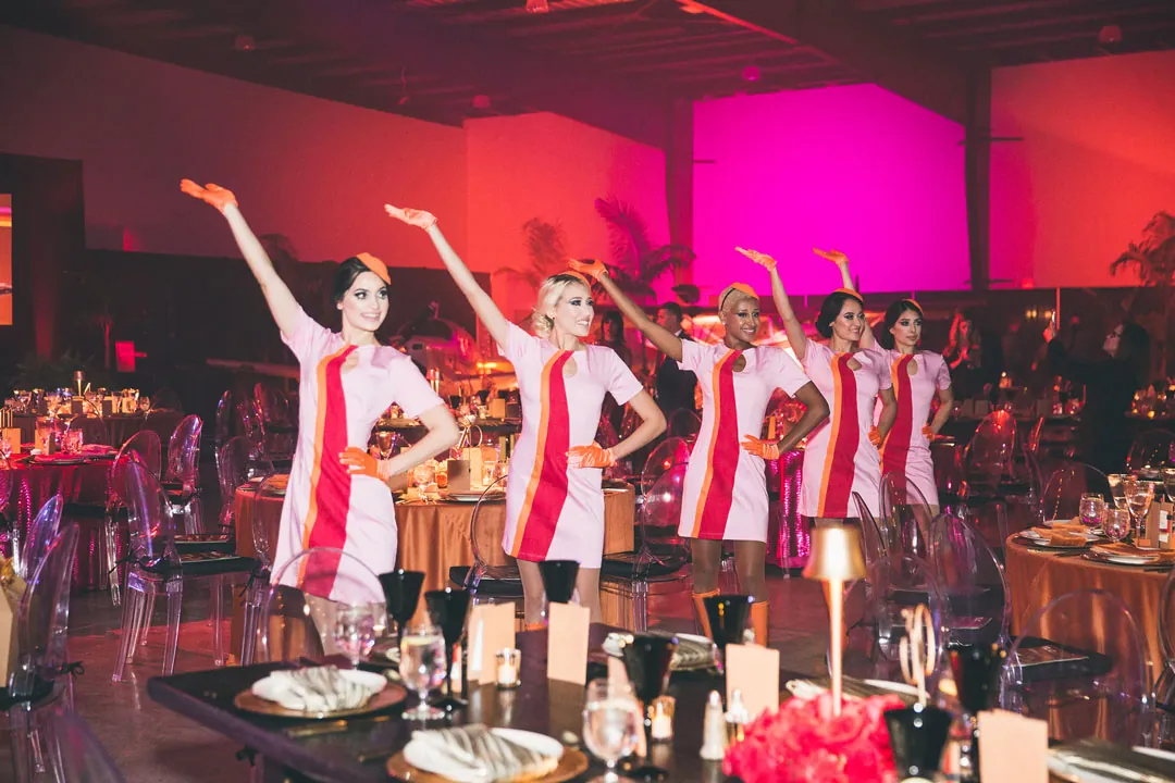 Five dancers in pink retro flight-attendant uniforms perform between dinner tables under pink uplighting at the gala