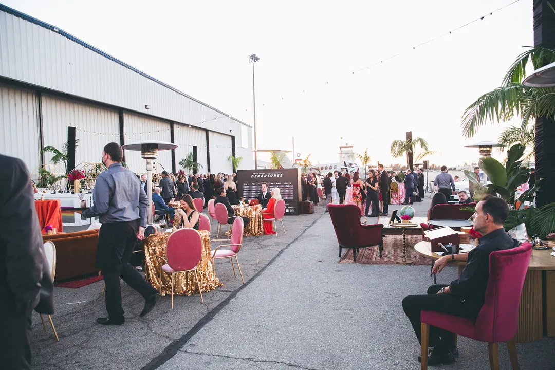 Cocktail hour on the tarmac at The Executive Hangar with sequin tables, velvet chairs, palms and a plane in the distance