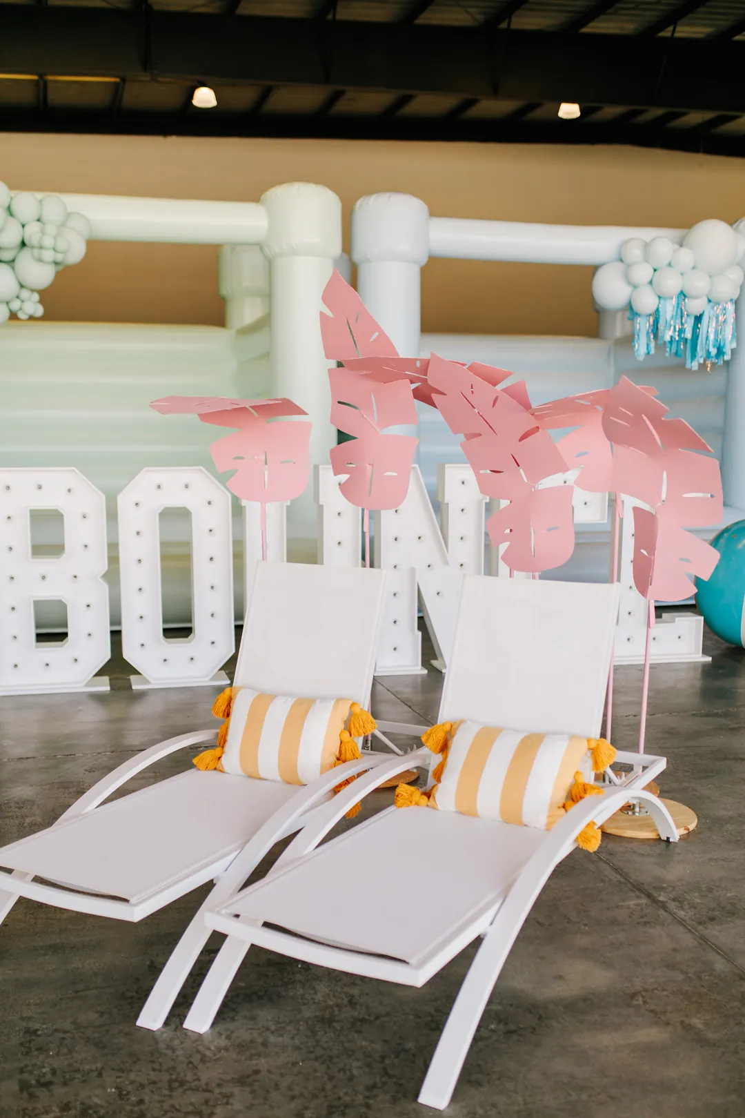 Two white lounge chairs with striped pillows under pink paper palm leaves by BOUNCE marquee letters and a mint bounce house