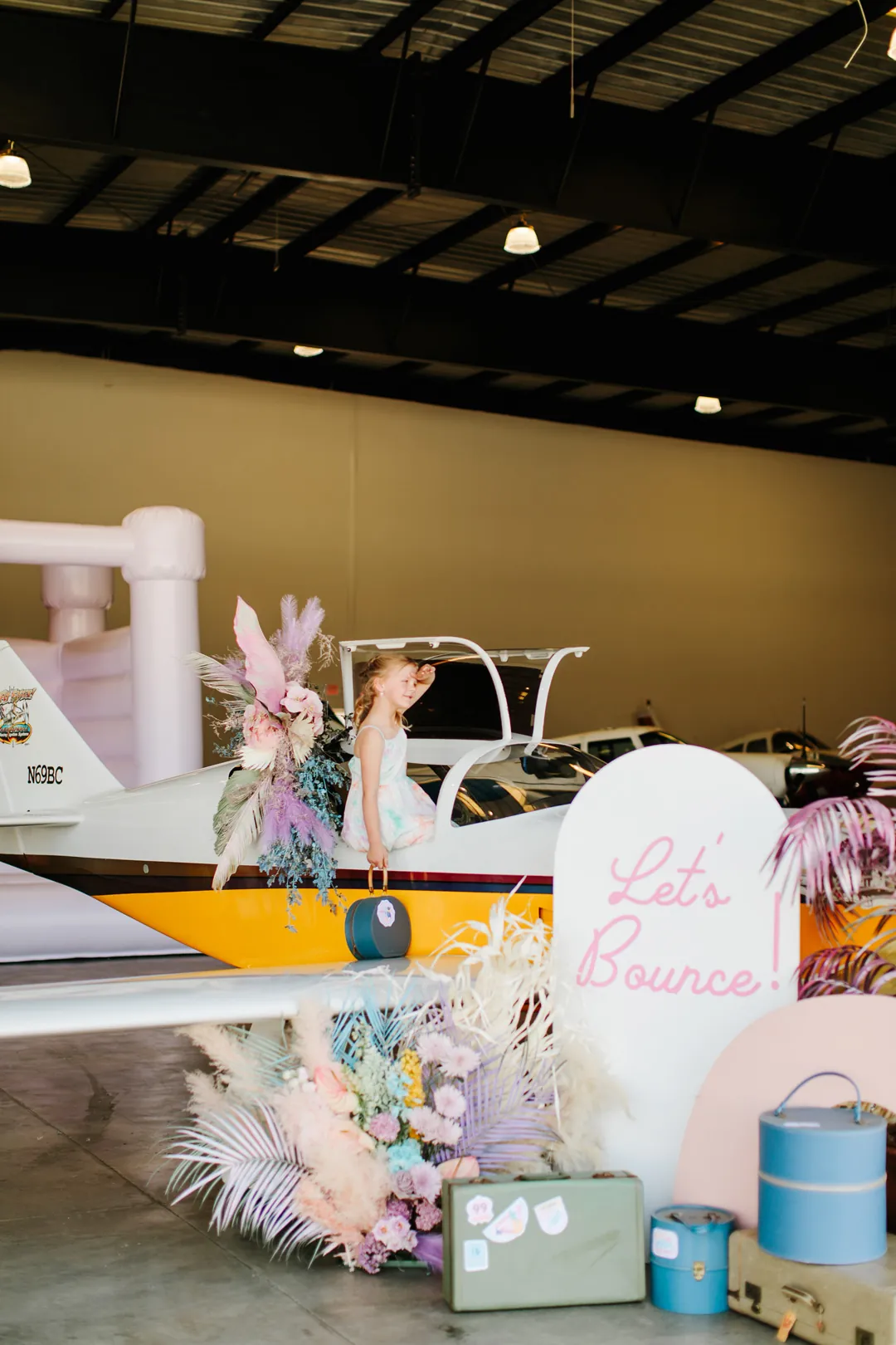 Little girl in the cockpit of a floral-decorated plane beside a Lets Bounce sign and vintage luggage at The Executive Hangar