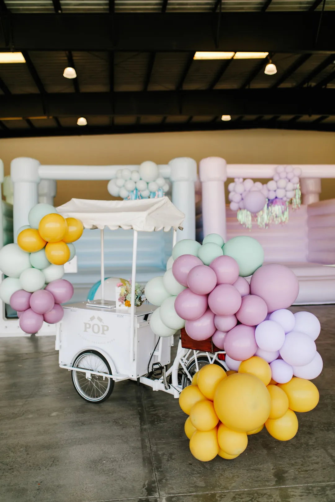 White POP popsicle tricycle cart wrapped in mint, lilac and mustard balloon garlands in front of pastel bounce houses