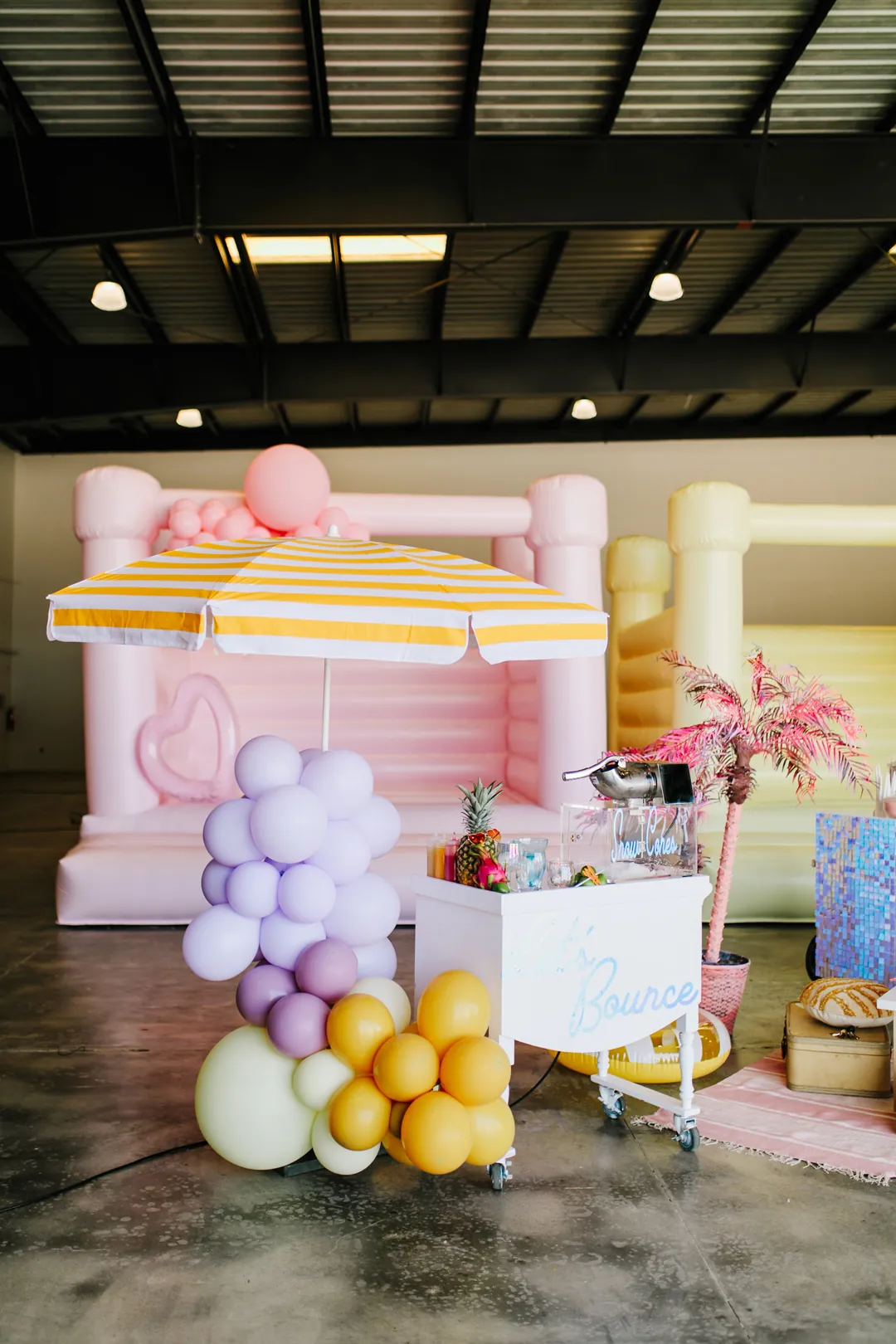 Lets Bounce snow cone cart under a yellow striped umbrella with lilac and mustard balloons in front of a pink bounce house