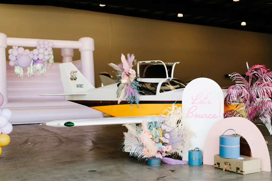 Small plane draped in pastel florals beside a Lets Bounce sign, lavender bounce house and vintage luggage at the hangar