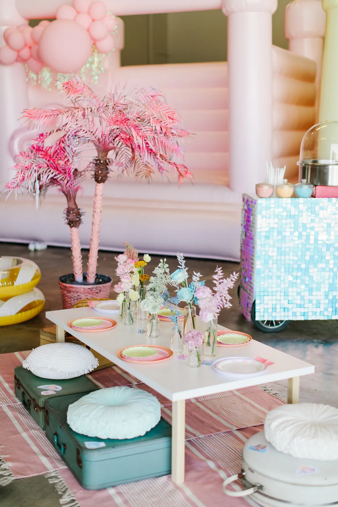 Low kids table with pastel plates, florals and suitcase seats by pink palm trees and a pink bounce house at the hangar