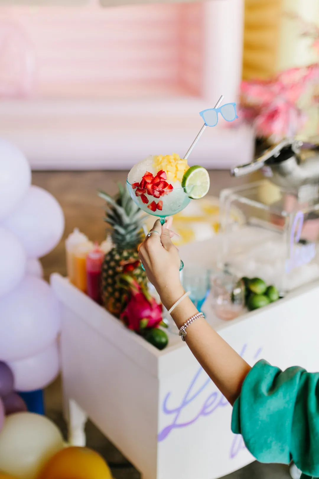 Guest holding a tropical snow cone topped with strawberries, pineapple and lime at a treat cart stacked with fresh fruit