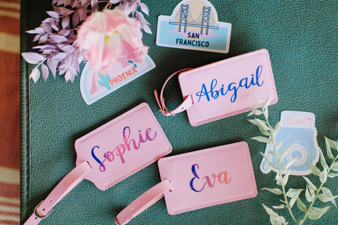 Three personalized pink luggage tags with hand-lettered names, travel stickers and greenery on a teal vintage suitcase